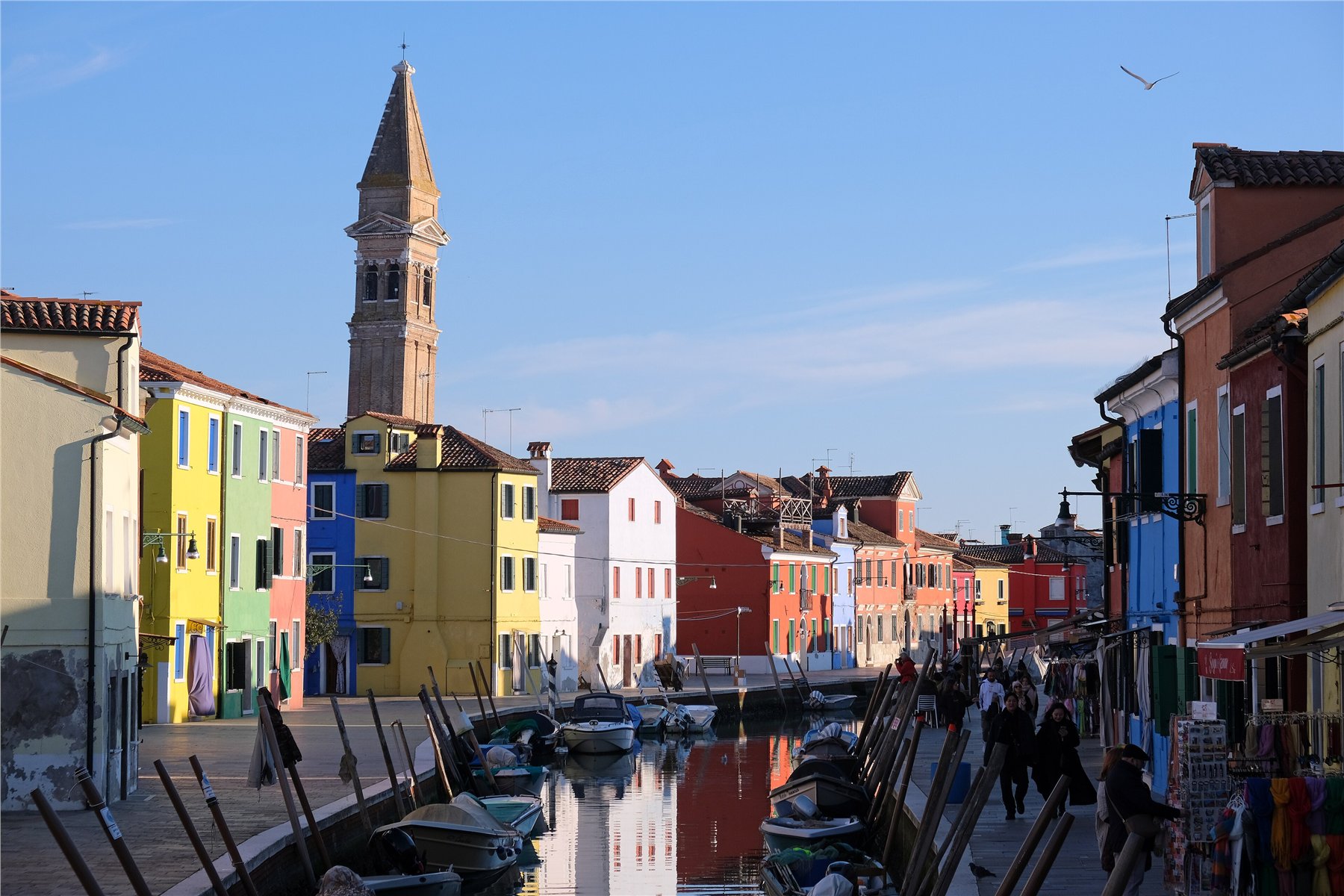 Rio Giudecca canal in Burano lined with saturated lemon-yellow, sage-green, coral-pink and cobalt-blue houses, the leaning bell tower of San Martino church in the distance, still water reflecting the whole street, a row of mooring poles in the foreground