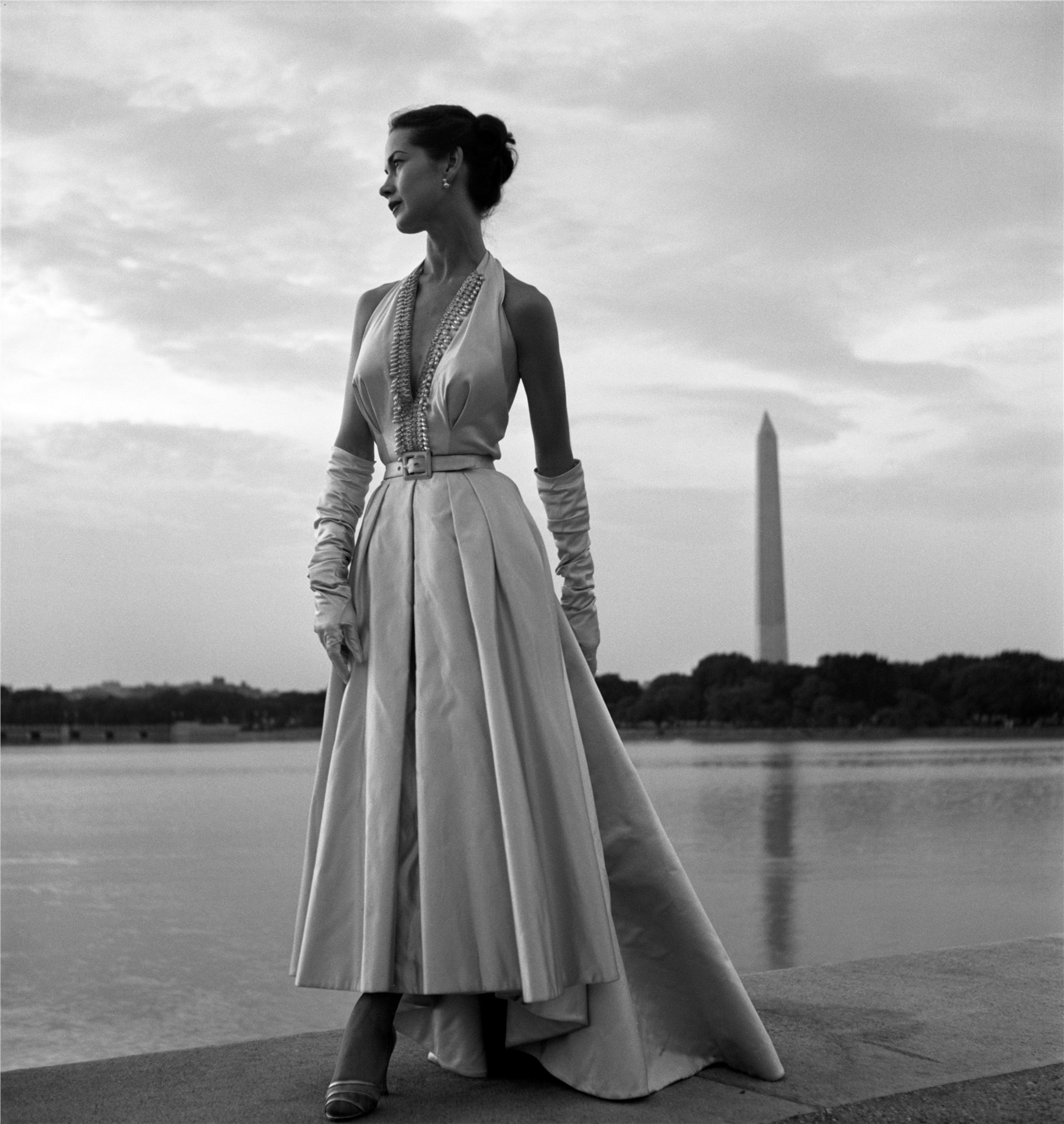 Black and white photograph of a model in a beaded halter-neck evening gown and elbow-length gloves, standing in profile on the steps of the Tidal Basin in Washington D.C., with the Washington Monument reflected in the water behind her under an overcast sky