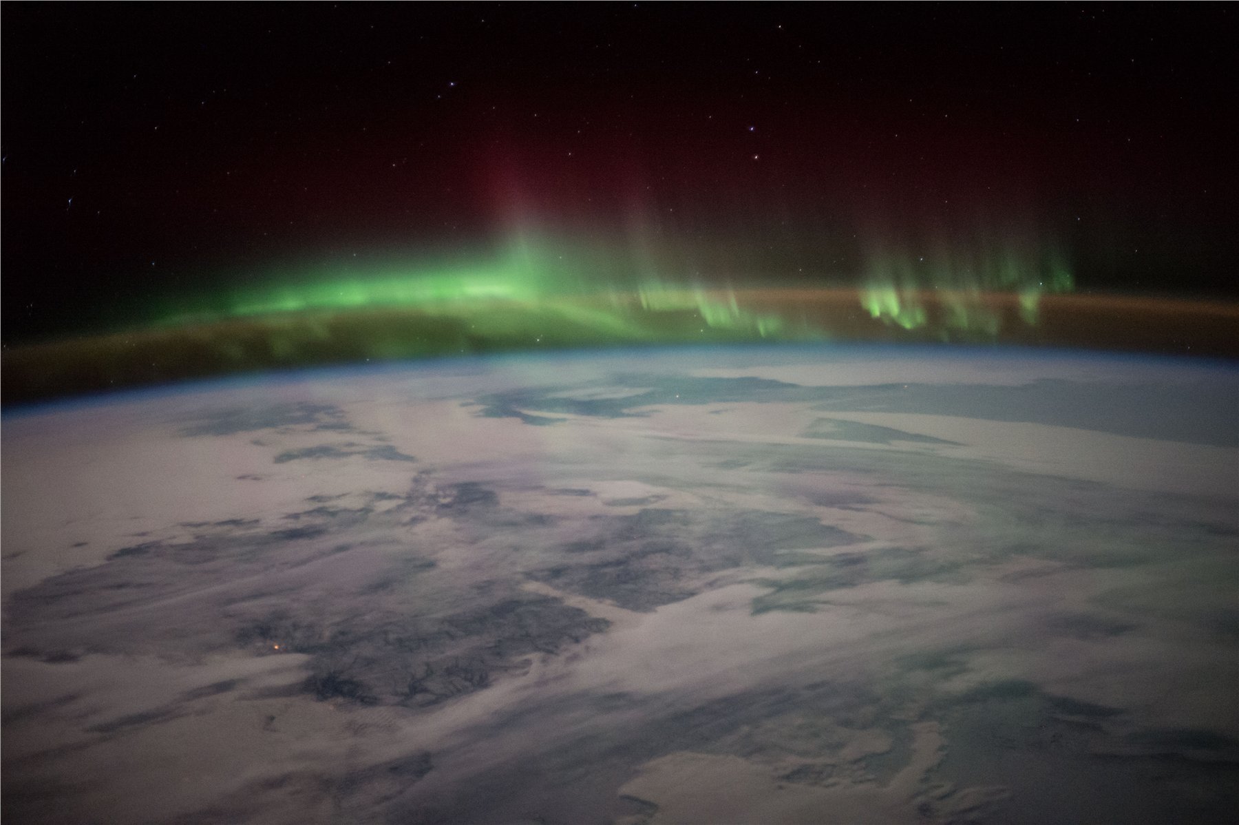 A view from the International Space Station of Earth's limb: snow-covered Canada below, a blue band of atmospheric glow above it, a wide green auroral curtain rippling above that, a faint crimson band of high-altitude aurora at the top, and a star-filled black sky beyond