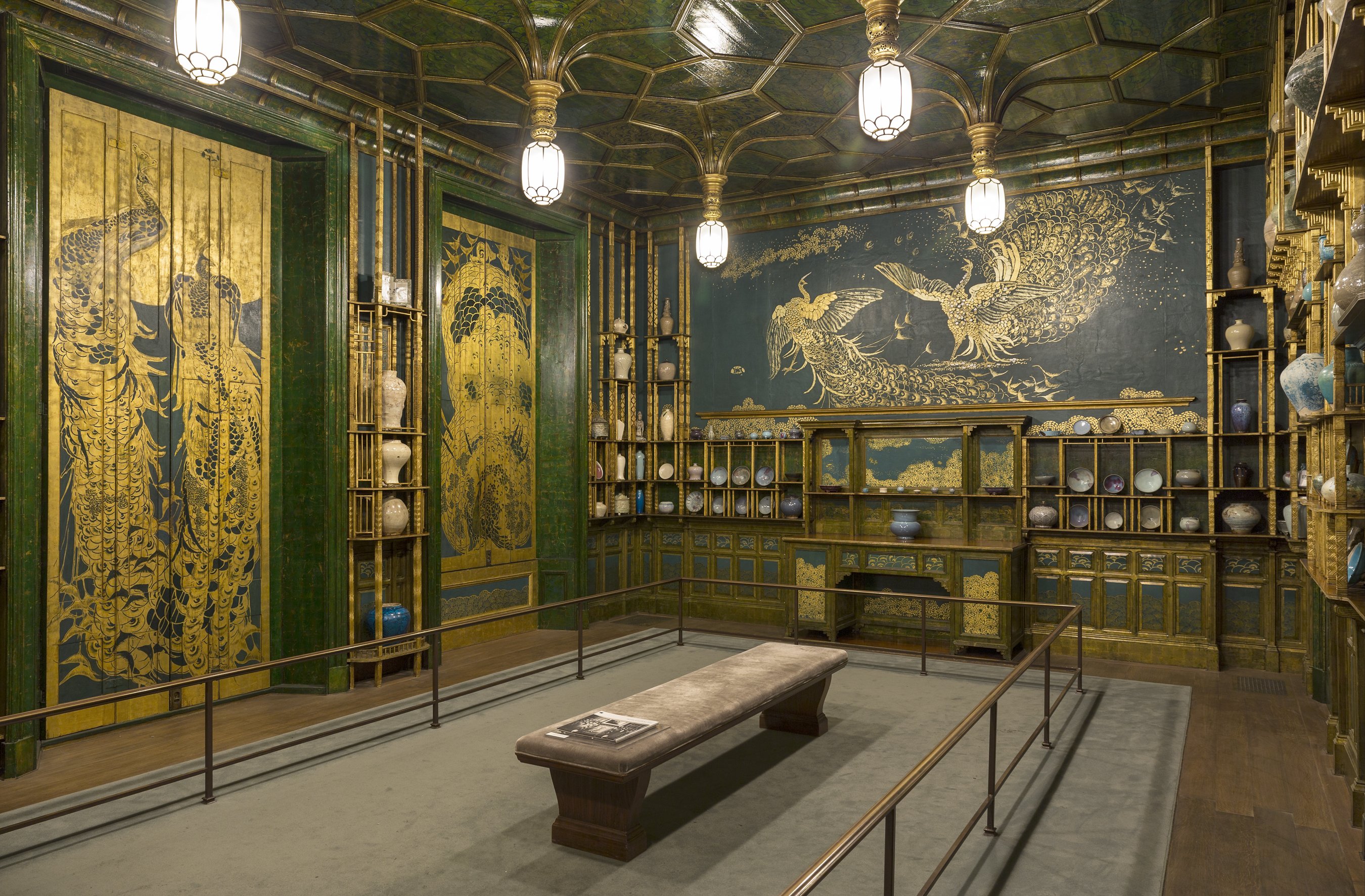 Interior of Whistler's Peacock Room: gilded shutters painted with peacocks on the left, a deep indigo mural of two facing golden peacocks on the far wall, gilt lattice shelving holding blue-and-white porcelain, and a honeycombed gilded ceiling with hanging lanterns