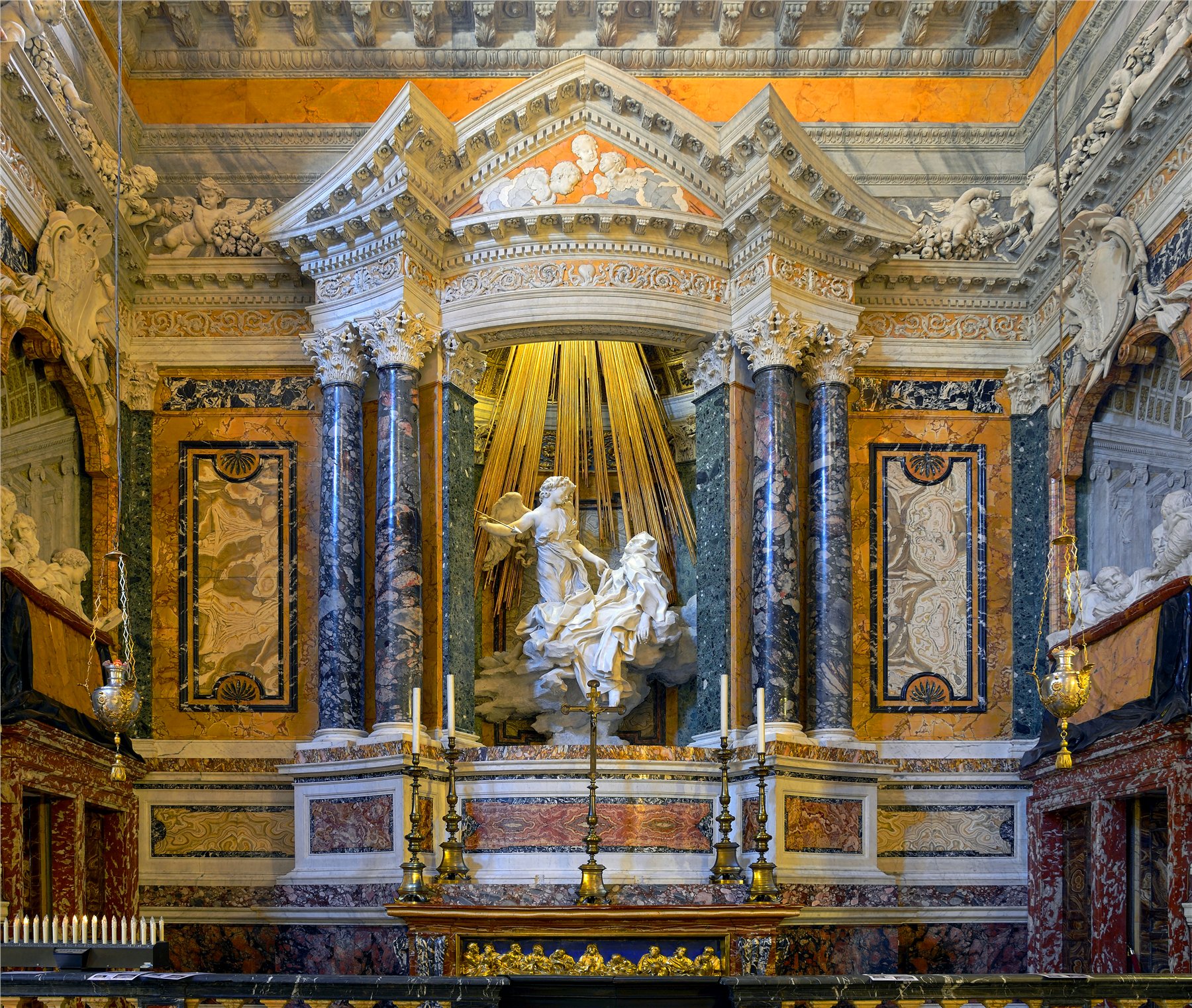 Interior of the Cornaro Chapel: a cascade of gilt bronze rays falling from a hidden upper window onto a white marble sculpture group of an angel and a swooning nun, floating on carved clouds within a frame of dark green marble columns and gold-veined ochre wall panels