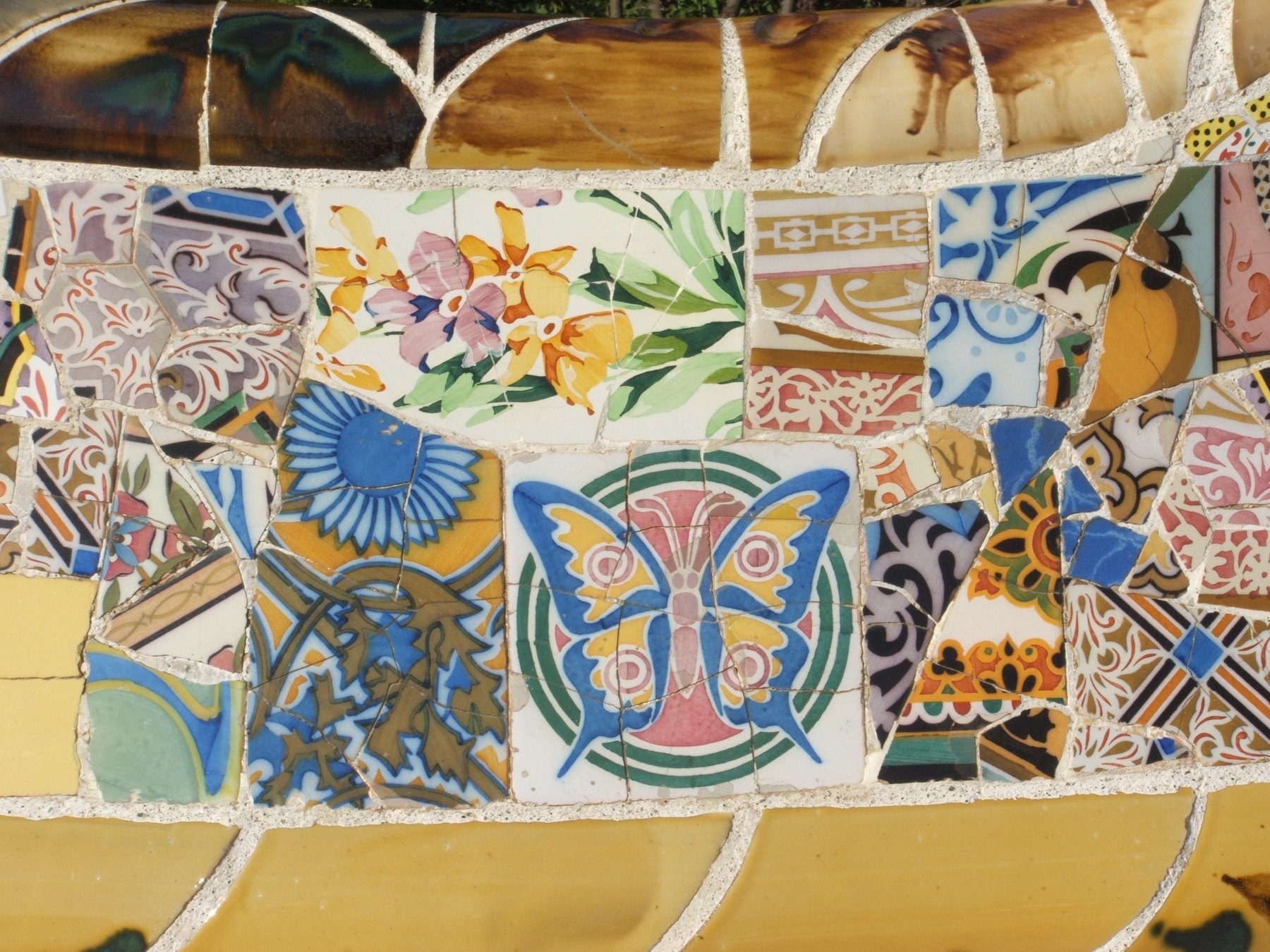 Close-up of Park Güell's serpentine bench: a hand-painted iris tile, a blue-and-gold butterfly medallion, a sunflower tile, and geometric ceramic fragments pieced together with white grout, bordered above by a glazed ochre rail