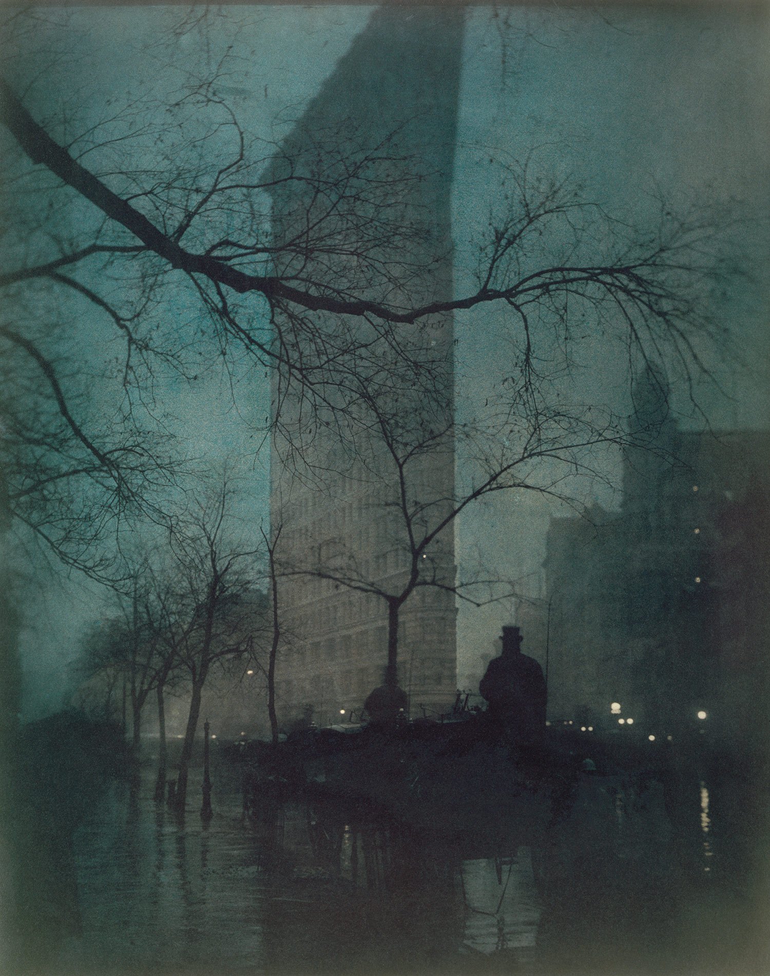 The Flatiron Building rising through rain and mist behind silhouetted branches, carriages, and pedestrians